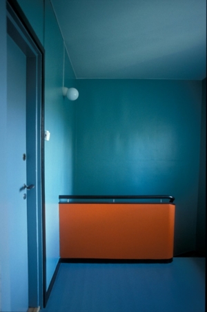 Trappegang med turkise vegger, blått gulv og dør, svarte dørkarmer og orange parti ved trappa.